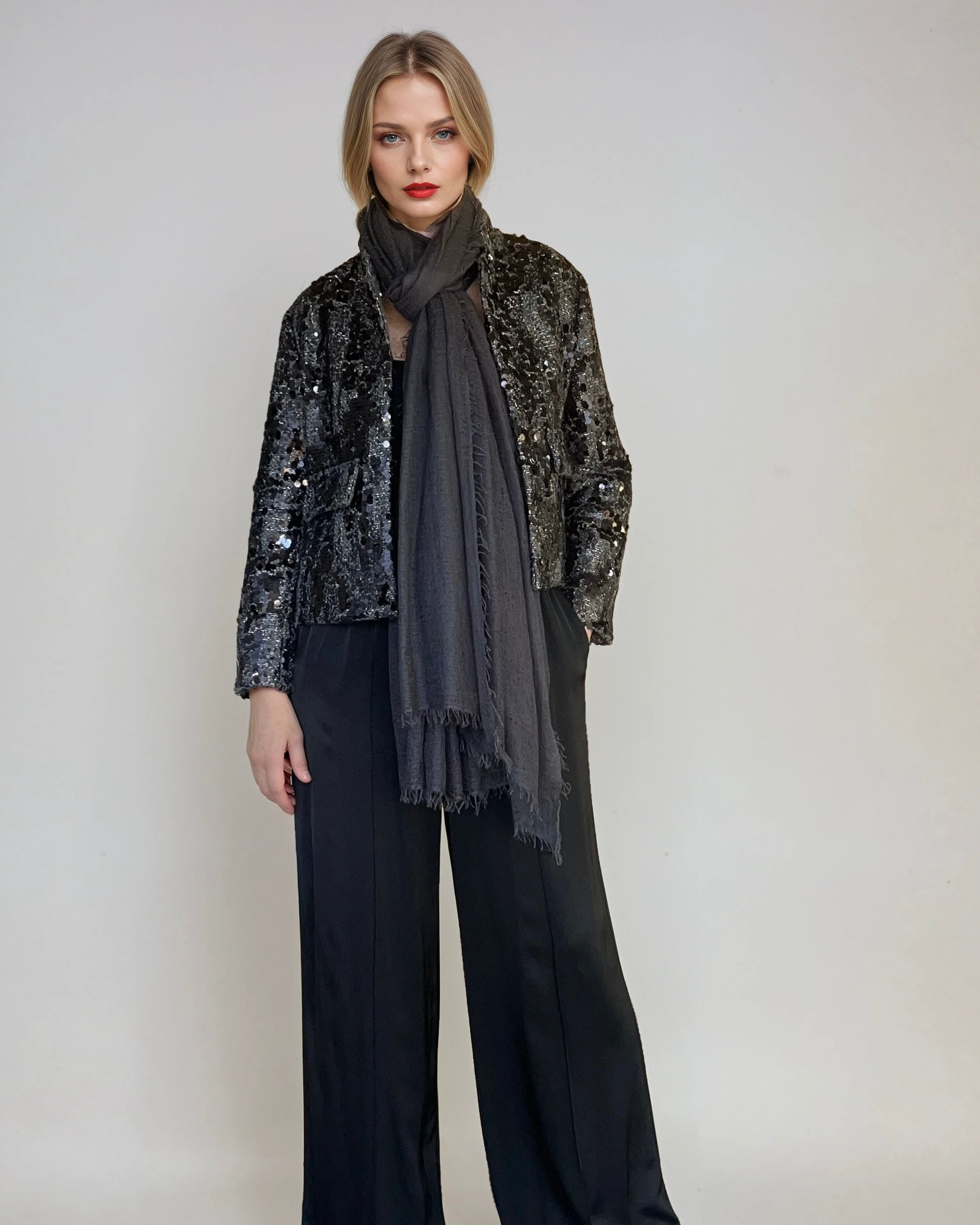 Woman wearing a sequined jacket, a charcoal extra large cashmere scarf, and black pants on a plain background