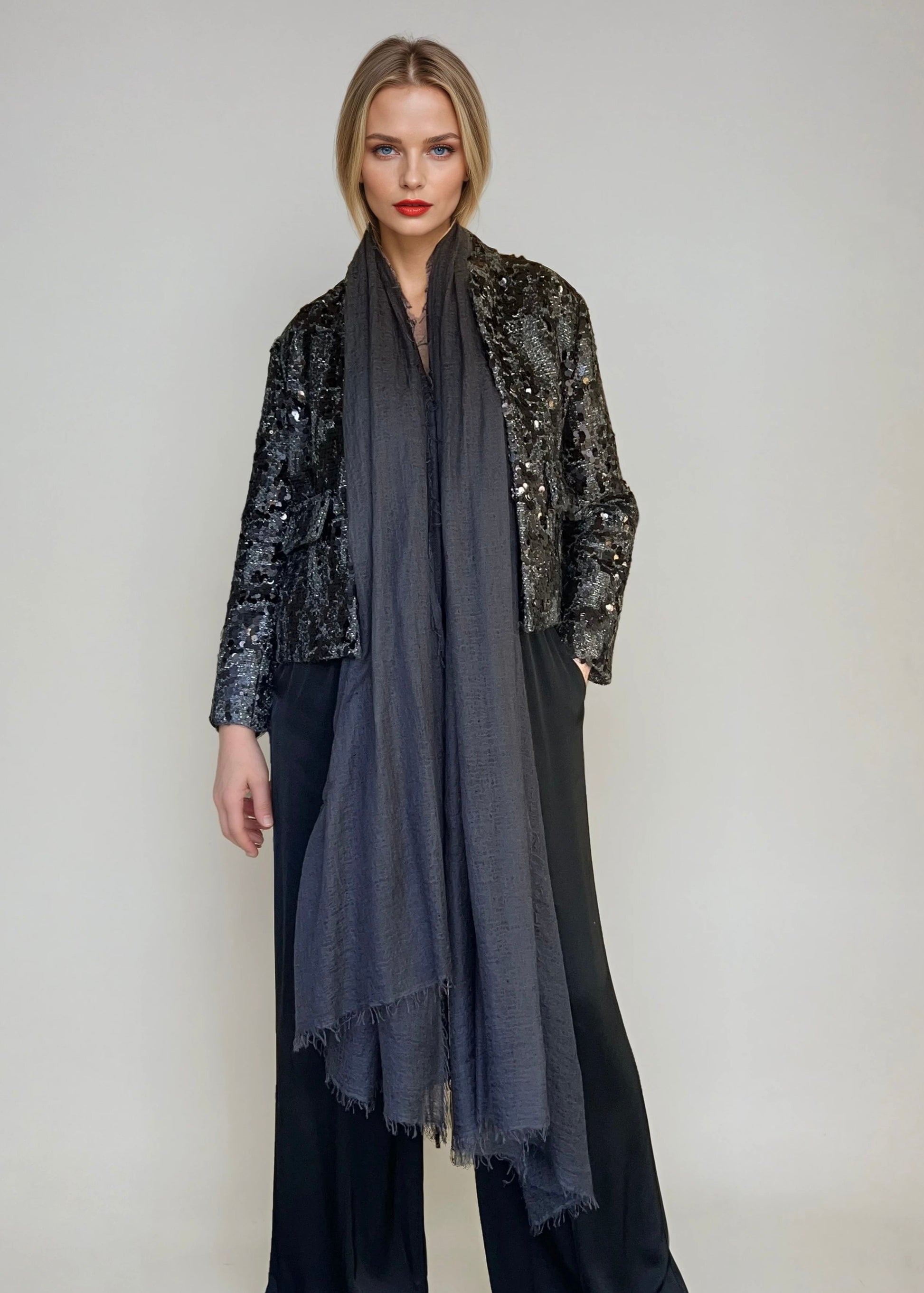 Woman wearing a black sequin jacket, extra large charcoal cashmere scarf, and black pants on a plain background