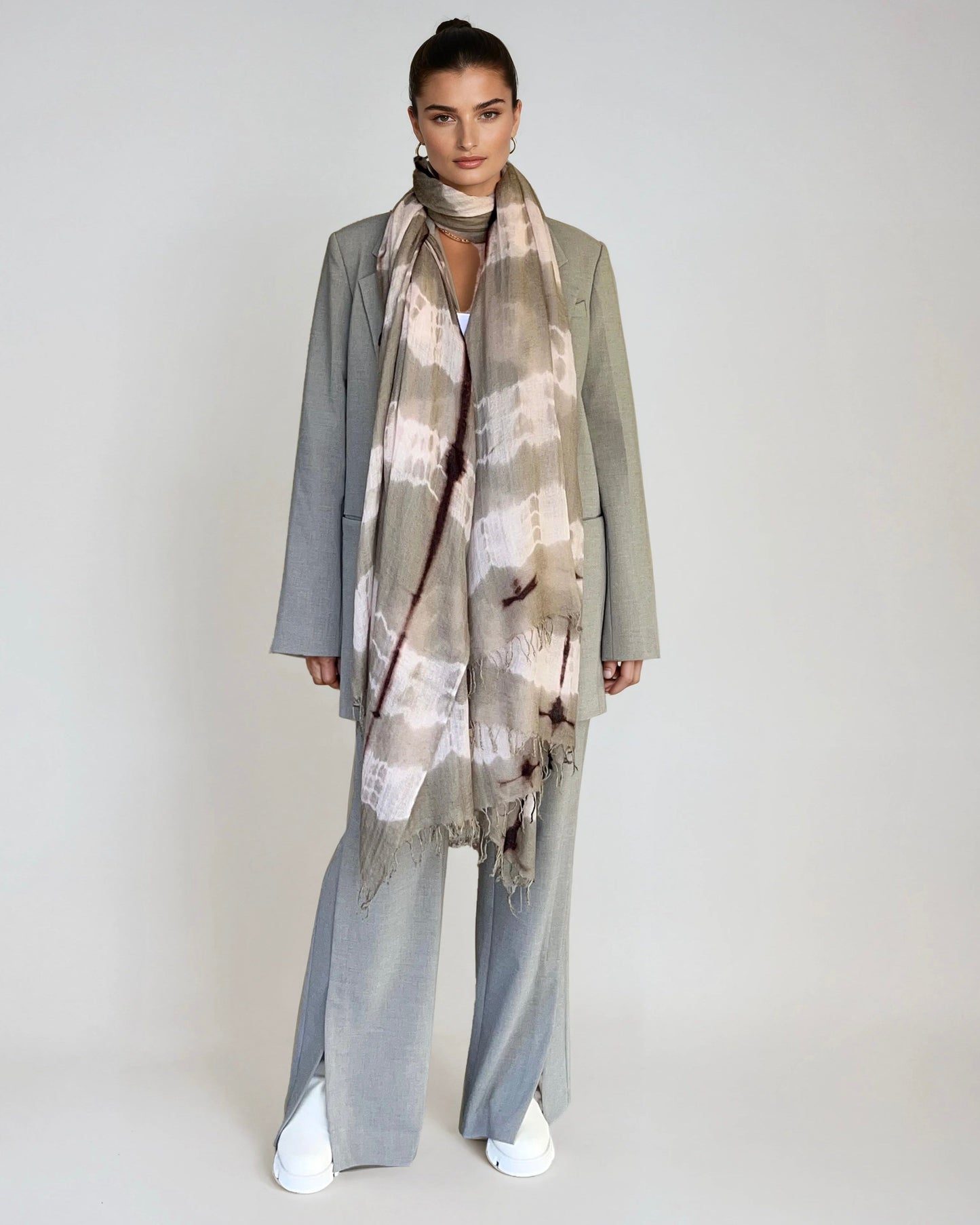 Woman wearing a long, light-colored scarf with a pattern against a plain background