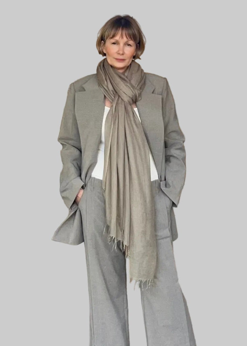 Person wearing a gray suit and scarf standing in front of a white wall.