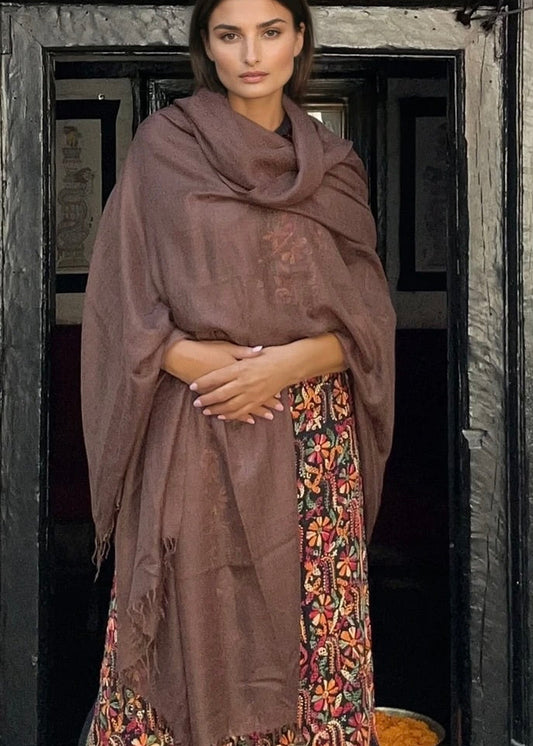 Woman wearing a brown shawl over a patterned dress standing in front of a dark wooden door.