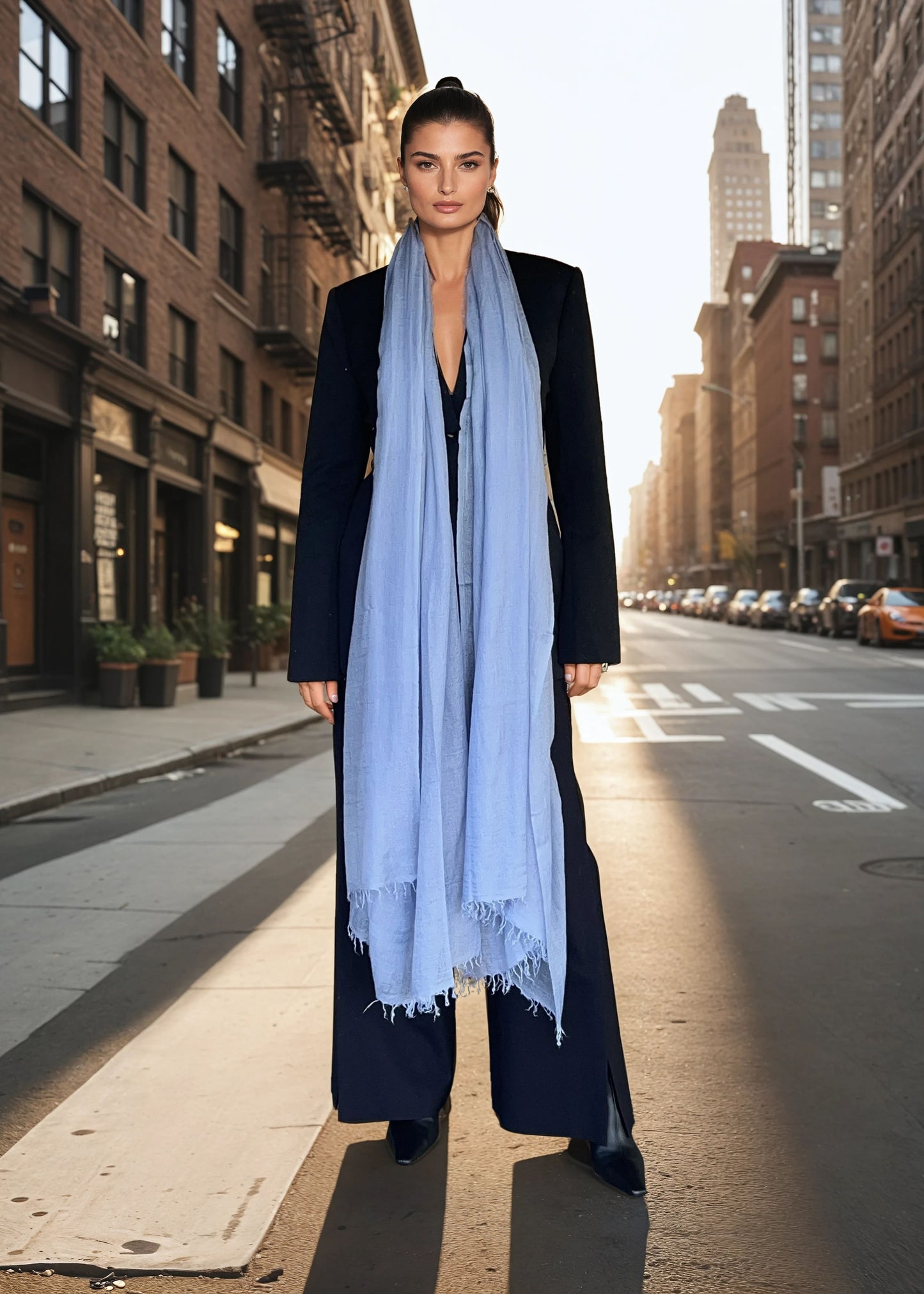 Woman wearing a long black coat and light blue scarf on a city street.